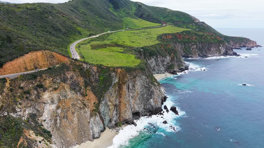 Drone photograph of Highway 1 winding along steep, layered cliffs in Big Sur, California, with a grassy plateau perched above the rugged shoreline. Turquoise water and rocky points extend along the coast below, with hazy mountains and headlands visible in the distance.