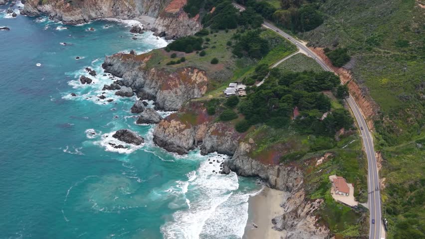 Drone photograph of Highway 1 curving along the rugged Big Sur coastline in California, with cars traveling past scattered trees and a small building near the roadside. Layered rocky cliffs and headlands extend along the shore, with turquoise water breaking against the rocks below.