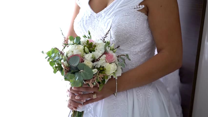 A woman dressed in a stunning white wedding gown holds a vibrant bouquet filled with greenery and delicate flowers. Light pours in, creating an atmosphere of excitement.