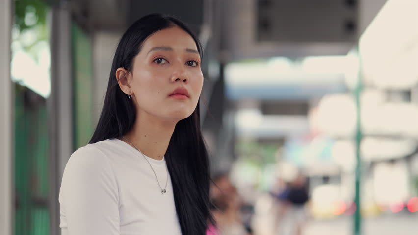 Medium shot of a stunning young Thai woman in a casual white long-sleeved shirt standing at a public transport station or walkway in Bangkok, looking contemplative then turning to smile at the camera. Portrait of modern Asian lifestyle and street fashion. #sstkFashion