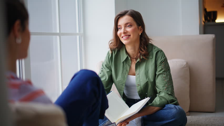 Carefree lady chatting friend holding book relaxing in modern apartment closeup. Smiling two women talking together sitting home floor. Happy besties sharing secrets discussing new at domestic leisure