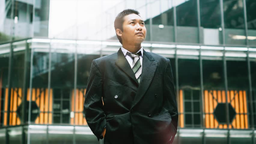 Low angle medium shot of a confident young Southeast Asian businessman in a sharp black suit standing outside a modern glass office building
