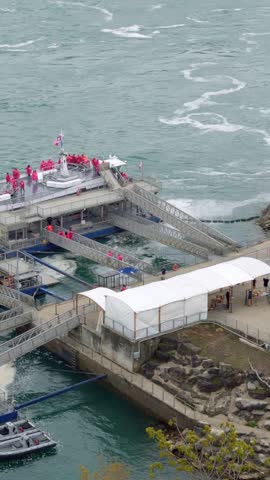 Aerial view of riverside pier with people in red jackets standing on platforms and stairs near white canopy, boats below and flowing water surrounding concrete waterfront structure.