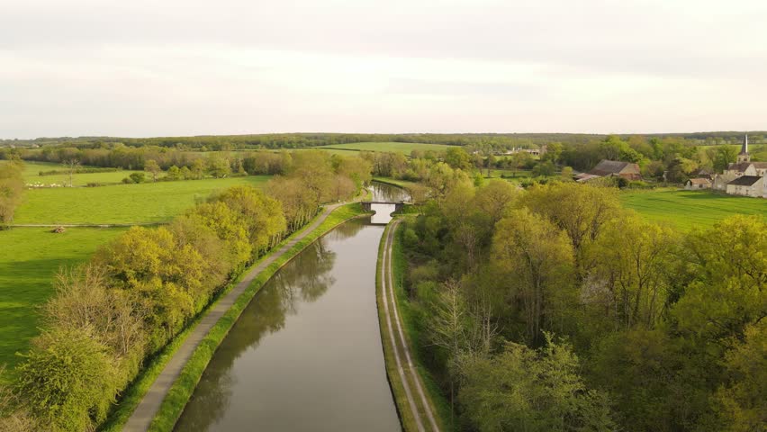 Aerial view of a calm canal surrounded by lush green trees and rural fields under a soft sky near Nevers, Bourgogne-Franche-Comte, France.