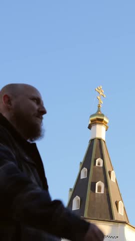 Bald bearded man gestures near church steeple, moving through frame against blue sky, with golden cross atop pointed tower and small windows on dark spire, emphasizing motion and architecture.