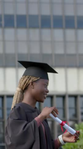 Vertical video: Turning diploma, grad in cap and gown tracking white object appearing above campus. Mortarboard, scroll, student, robe, outdoor, campus, vertical video