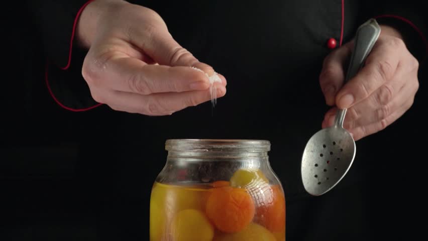 Chef hands in a black uniform sprinkle seasoning over a jar filled with vibrant preserved fruits, highlighting the careful attention to flavor enhancement in a dimly lit kitchen environment.