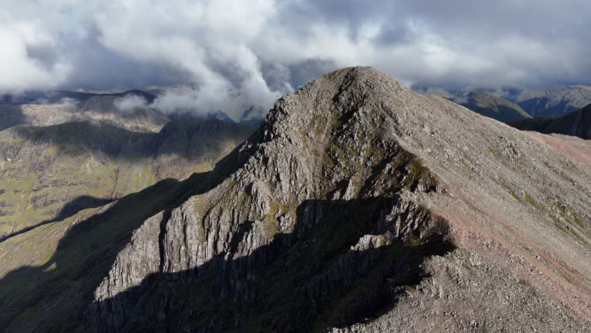 A sweeping perspective glides past this craggy, sun-drenched summit. The dramatic, rocky massif dominates the wilderness, framed by shifting clouds above the vast, ancient British landscape.