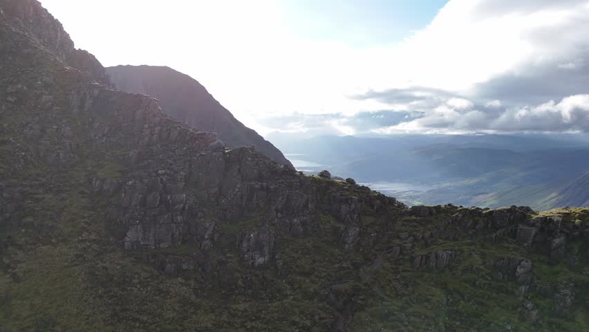 An aerial shot captures a solitary adventurer navigating a rugged mountain crest. The expansive vista reveals dramatic crags and distant valleys under a bright, cloudy sky. Part of the Three Sisters.