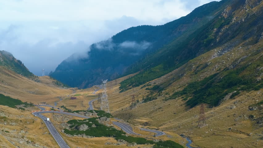 Clouds passing over mountain road with winding curves. Winding roads traverse a mountain valley, with cars navigating the curves. Low clouds drift over the hills, creating a picturesque landscape