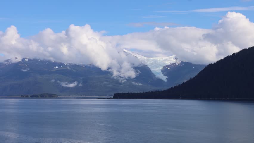 Aerial view of the Tracy Arm-Fords Terror Wilderness featuring a glacier nestled between forested mountains and clouds over the calm water in Juneau, Alaska, United States.
