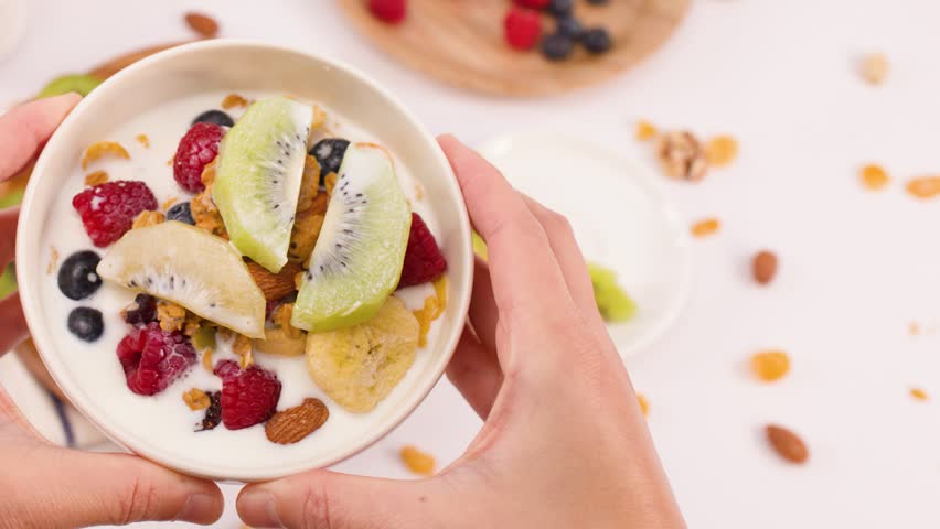 Top-down view of hands setting a colorful yogurt bowl onto a bright, ingredient-filled table.
