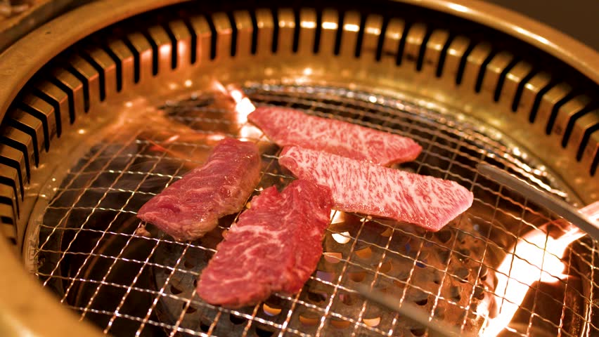 Close-up shot of marbled beef strips cooking on a circular metal grill with flickering flames.