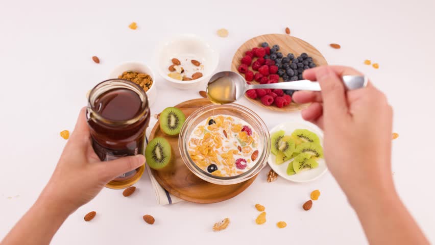 Overhead shot of hands pouring honey into a breakfast bowl with milk and fruit.