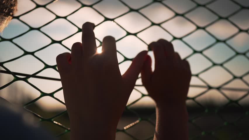 Two hands grip chain link fence at sunset outdoors. Person holds metal fence with sunlight between fingers. Hands touch fence during golden hour. Sunset shines through chain link and fingers.