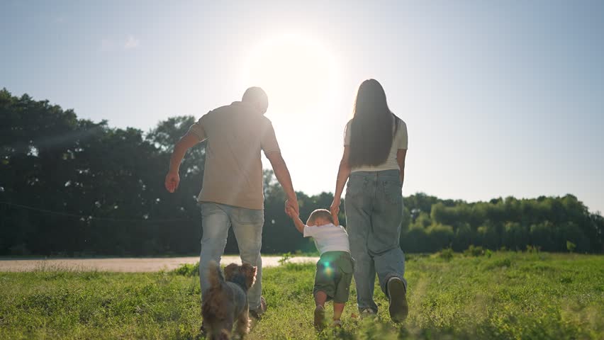 Parents and child walking hand in hand. Dog running nearby. Summer grass, green field, blue sky. Family walking, holding, walking again. Dog and boy happy. Family enjoying summer nature together.