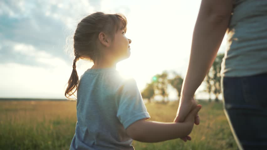 Mother and child walking together outdoors. Young daughter gently holds mother hand at warm sunset. Little child feels calm. Family keeps walking across green summer park. Parent looks joyful.