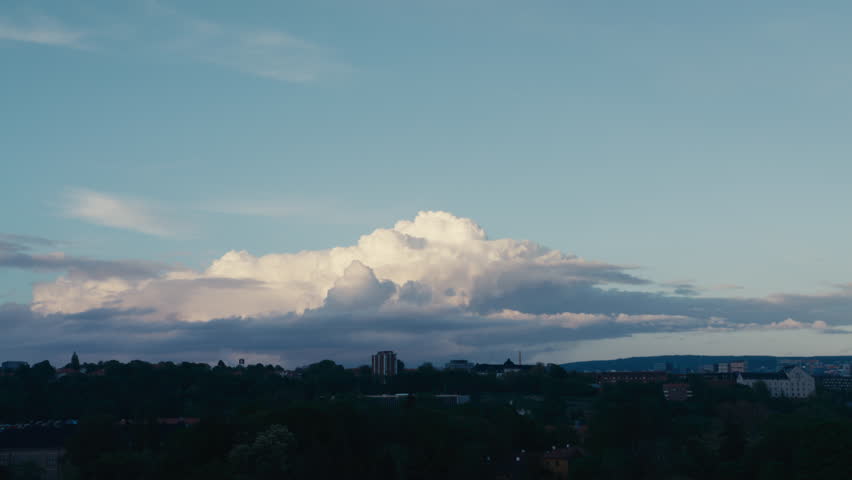 Large cumulus clouds glow in the evening light above a dark Norwegian town skyline with distant hills and soft blue sky.