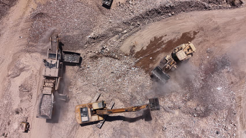 Overhead view of an active mining quarry with heavy machinery including excavators and dump trucks moving rocks and debris across a dusty open-pit site