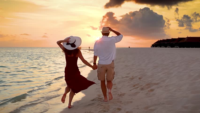 Slow motion view of a beautiful couple holding hands and running down a tropical beach during sunset time in the Maldives islands