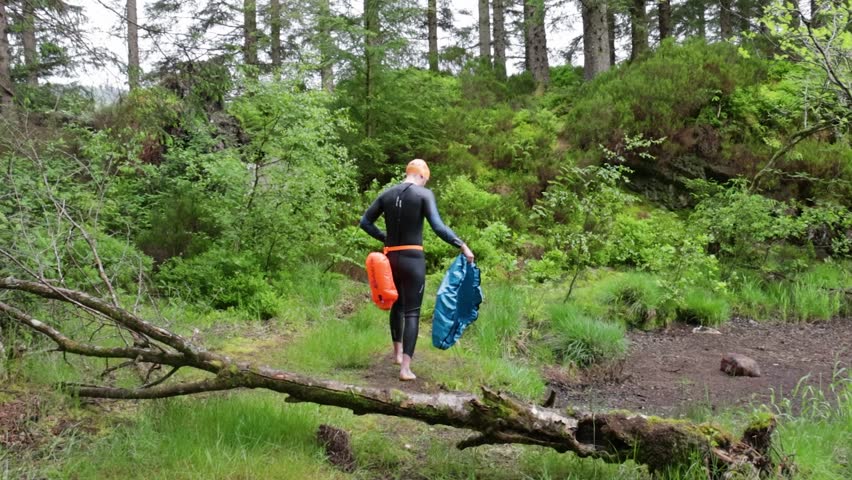 Male wetsuit swimmer heads to a forest lake, carrying an orange safety tow float on a muddy trail by the water’s edge.