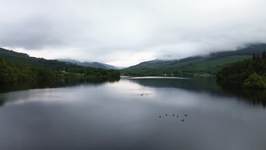 Loch Ard rising drone shot reveals broad freshwater lake framed by pine woodland and rolling Highlands hills under low cloud, ideal for travel, nature, and landscape use.