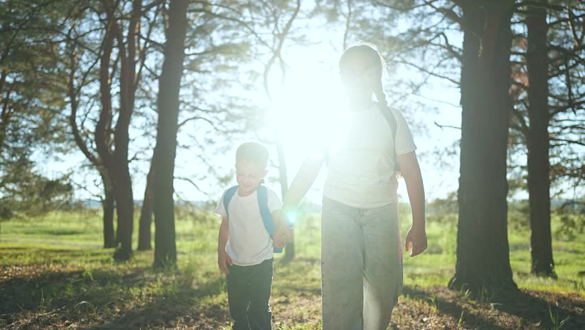 Sister boy hold hands hike. Brother walk next to sister with blue backpack. Trees forest around child. Girl smile walk with boy. Happy child explore forest together. Walk hike under bright trees.
