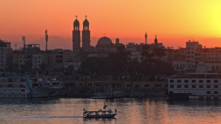 A vibrant golden sunset illuminates the East Bank skyline of Luxor, highlighting the prominent twin bell towers of Saint Mary Coptic Orthodox Church overlooking a cruising riverboat on the Nile.