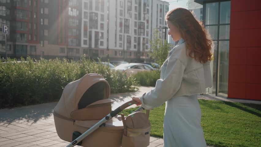 Side view tracking shot of a young redhead mother walking with a baby stroller on a sunny day. A woman pushing a pram along a sidewalk in a modern residential district with apartment buildings