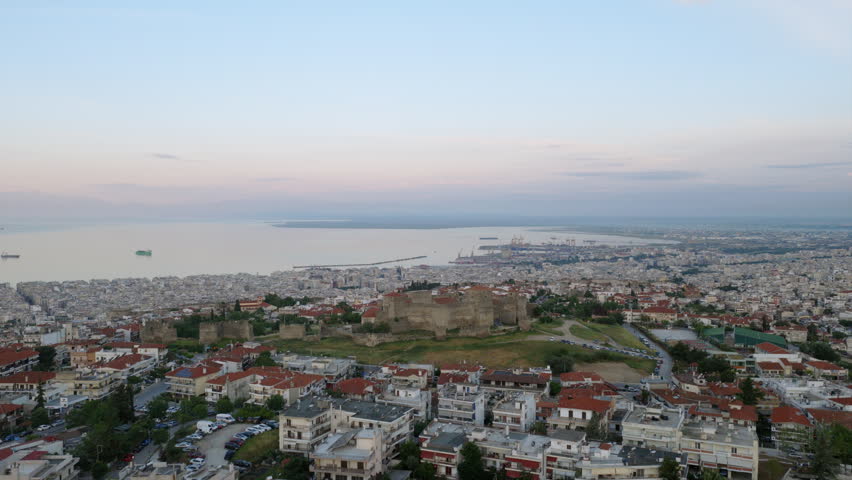 Sunrise aerial drone view of the Heptapyrgion fortress walls in Thessaloniki, Greece, with the city and Thermaic Gulf stretching toward the horizon.