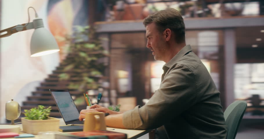 360 Degree. Caucasian Man Using Laptop Computer While Working in Brick Loft Office, Comparing Printed Spreadsheets Against Digital Analytics on Screen. Orbital Cinematic Arc Tracking Shot.