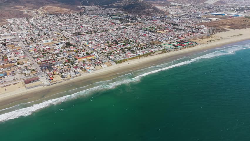 Drone photograph of a coastal town in Baja California, Mexico, showing a dense grid of colorful buildings stretching along a wide sandy beach. Ocean waves break gently along the shoreline, with dry, rolling hills rising behind the town under a clear sky.