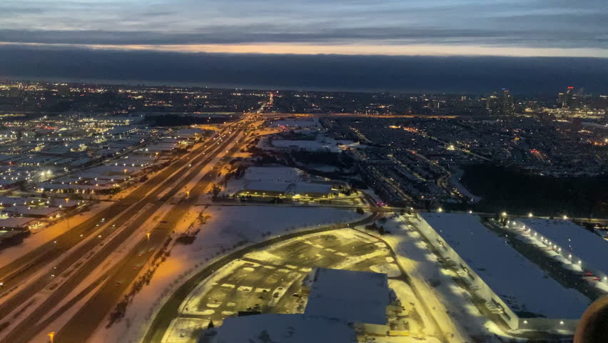 Aerial approach view from plane of Toronto at dusk in winter. Orange lit highway and snow covered industrial buildings, city lights glowing against sunset sky.