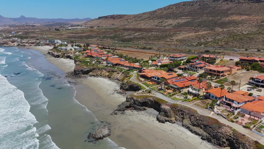 Drone photograph of a hillside beach community in Baja California, Mexico, featuring red-roofed homes lined along a sandy beach and rocky shoreline. Ocean waves roll in along the coast, with a sparse, dry landscape and distant mountains extending behind the neighborhood.