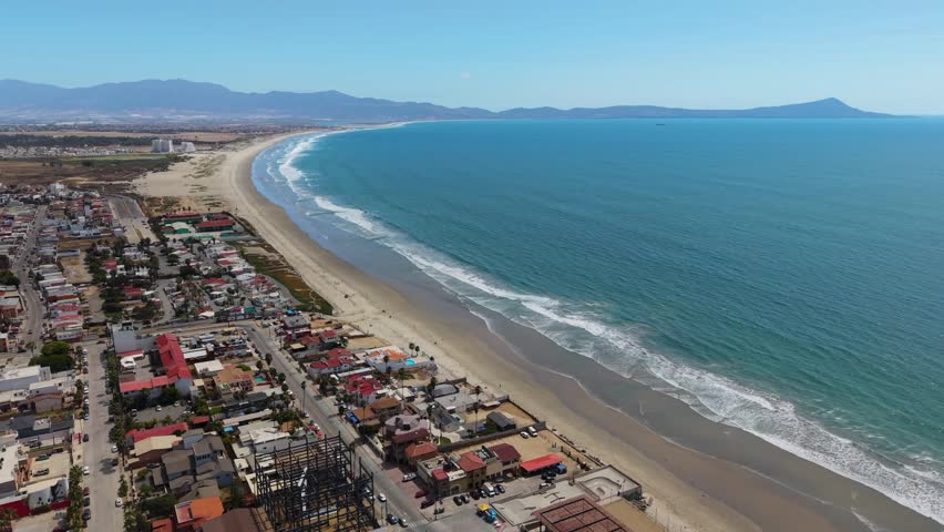 Drone photograph of a coastal town in Baja California, Mexico, with a long curving sandy beach stretching along the shoreline. Ocean waves roll gently onto the sand, with rows of homes and buildings lining the town and mountains rising in the hazy distance.