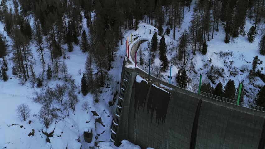 Oden Dam, Maloja, Switzerland—dolly shot with tilt-down over a snowy alpine dam, revealing the curved concrete wall, forested slopes and frozen winter valley