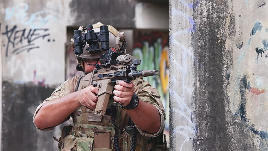 Armed tactical operator in camouflage gear poses inside a graffiti-covered industrial structure with reflective wet flooring.