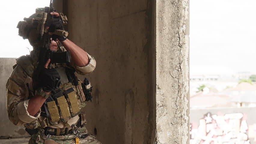 Armed tactical operator in camouflage gear poses inside a graffiti-covered industrial structure with reflective wet flooring.