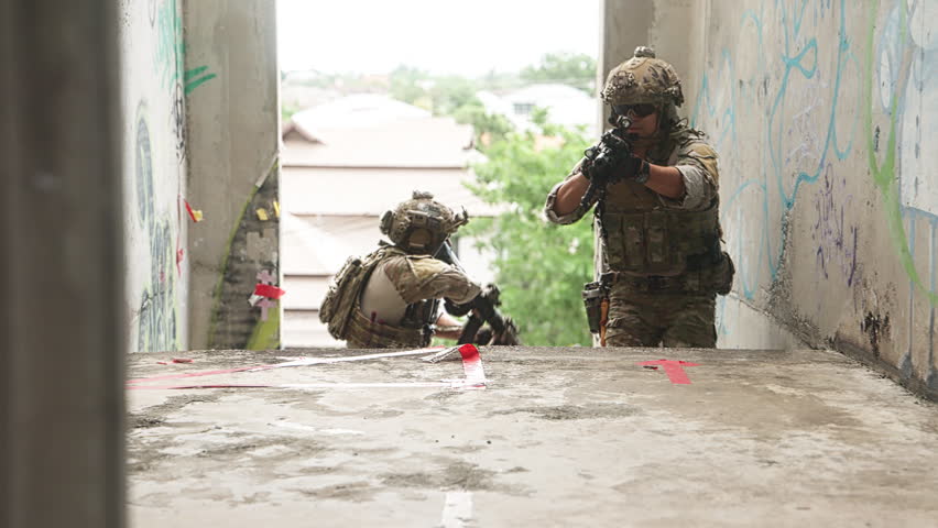 Two soldiers in camouflage tactical gear inside a concrete building with graffiti. One crouches while aiming forward, and the other stands aiming in the same direction. The scene highlights teamwork, discipline, and close quarters combat readiness in a realistic urban training environment. Suitable for defense, security, and tactical publications.