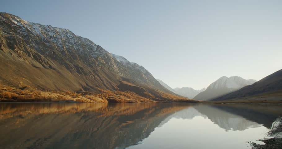Serene mountain lake reflects rugged peaks and golden forest in peaceful alpine wilderness landscape. Wide panorama shows calm water, snow dusted mountains, autumn trees, clear sky, rocky shoreline
