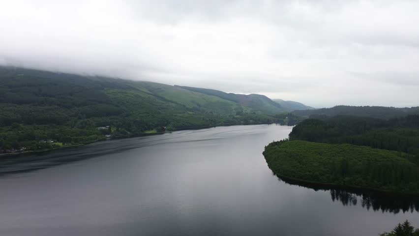 Loch Ard drone pullback reveals broad freshwater loch framed by dark conifer forest and rolling Highland hills under low cloud and grey skies, creating moody Scottish landscape scenery.