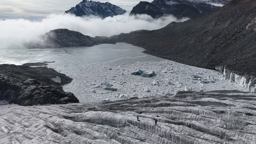 Aerial flyover over two hikers on the ice of Fellaria glacier in the Italian Alps on a grey autumn day crossing in between the glacial crevasses with an inversion and glacial lake in background