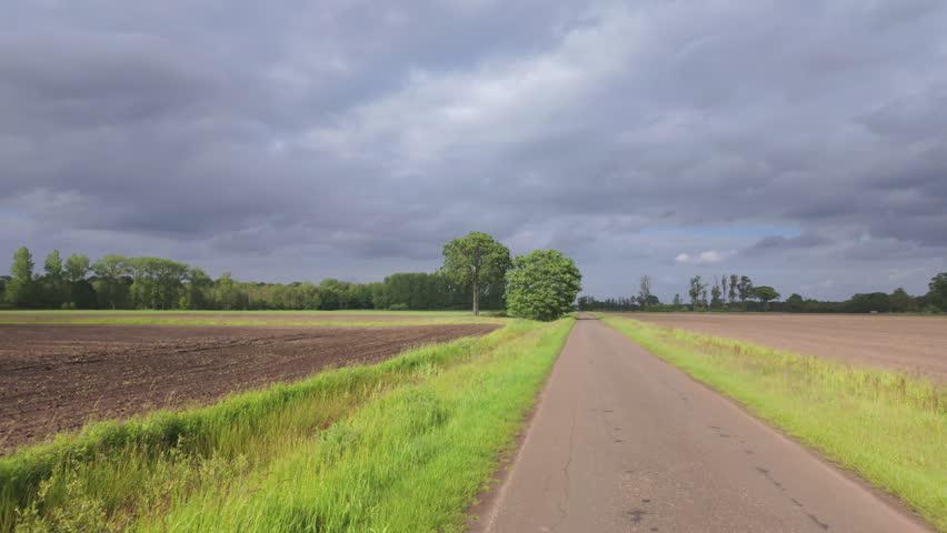Cinematic forward motion along a quiet country road in the Belgian Campine, passing farm fields under a dramatic, stormy sky.