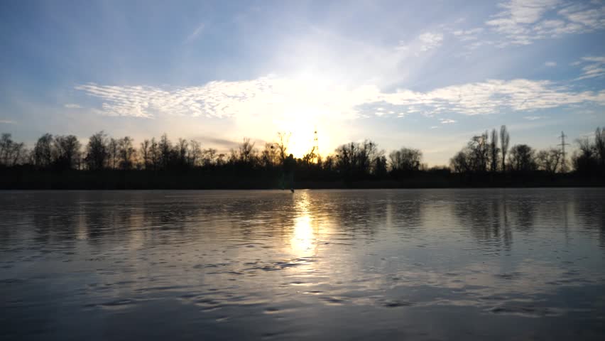 Young man skating on frozen river during sunset time. Guy shod in figure skates sliding on icy lake during training. Shining sun is reflecting on surface of ice. Active leisure at winter. Slow motion