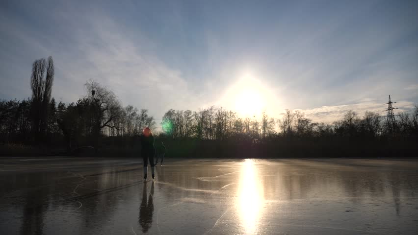 Two young people in figure skates sliding on ice during sunset. Man and woman skating on frozen river. Shining sun is reflecting on surface of ice. Couple have active leisure at winter. Slow motion