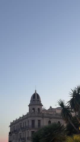 Vertical video of a historic domed building framed by palm trees in Tarragona, Catalonia, Spain. The elegant architecture stands against a clear evening sky, creating a peaceful Mediterranean cityscape at dusk. Travel destination, cultural heritage, historic architecture, urban tourism, and European landmark concept.