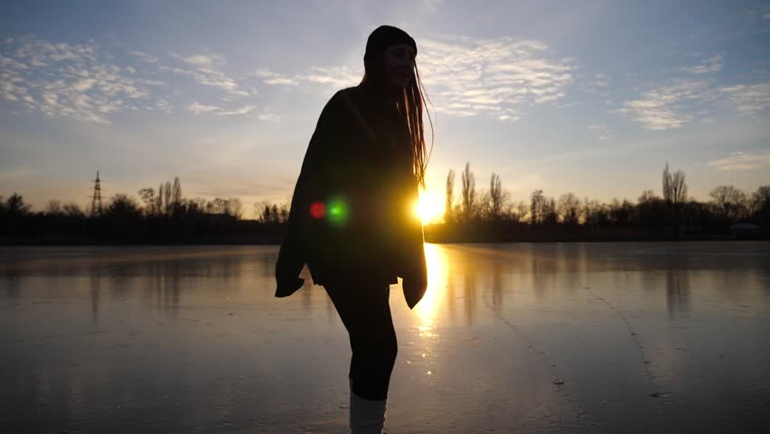 Female skater shod in figure skates sliding on ice surface during beautiful sunset. Sportswoman skating on frozen river. Silhouette of woman training at lake. Shining sun reflecting on surface of ice