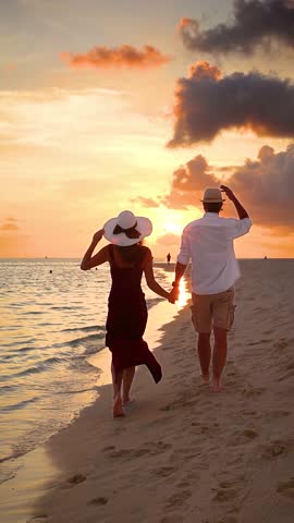 Rear view of a romantic couple on vacations holding hands and walking down a tropical beach during sunset time