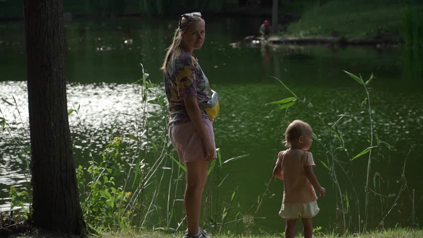a girl with a baby approach the edge of the water of a beautiful green city lake in a park, looking at fish and birds, tall grass and reeds along the shore, a warm summer day