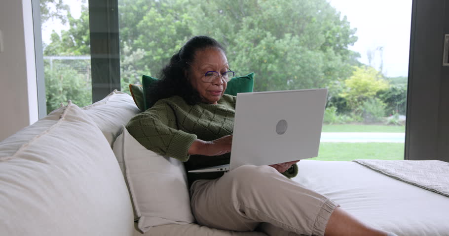Senior African American woman sitting on sofa in green knit using laptop shifting glancing outside. Homeoffice, typing, couch, window, garden, foliage, daylight